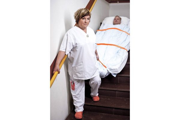 Eine Frau in weißen Arbeitsklamotten schiebt eine Matratze mithilfe eines Evakuierungstuchs eine Treppe hinunter. Die Matratze ist weiß und mit orangen Gurten befestigt.
